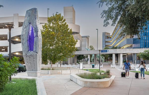 Plume, a metallic and purple obelisk, stands at the Austin airport with a parking garage in the background