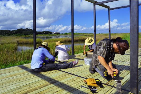 Several people kneeling and sitting as they work on a wooden platform that is surrounded by glass walls that overlook a marshy area and a blue sky