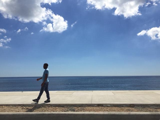 A person walking along a sidewalk next to a blue sky and a body of water on the horizon