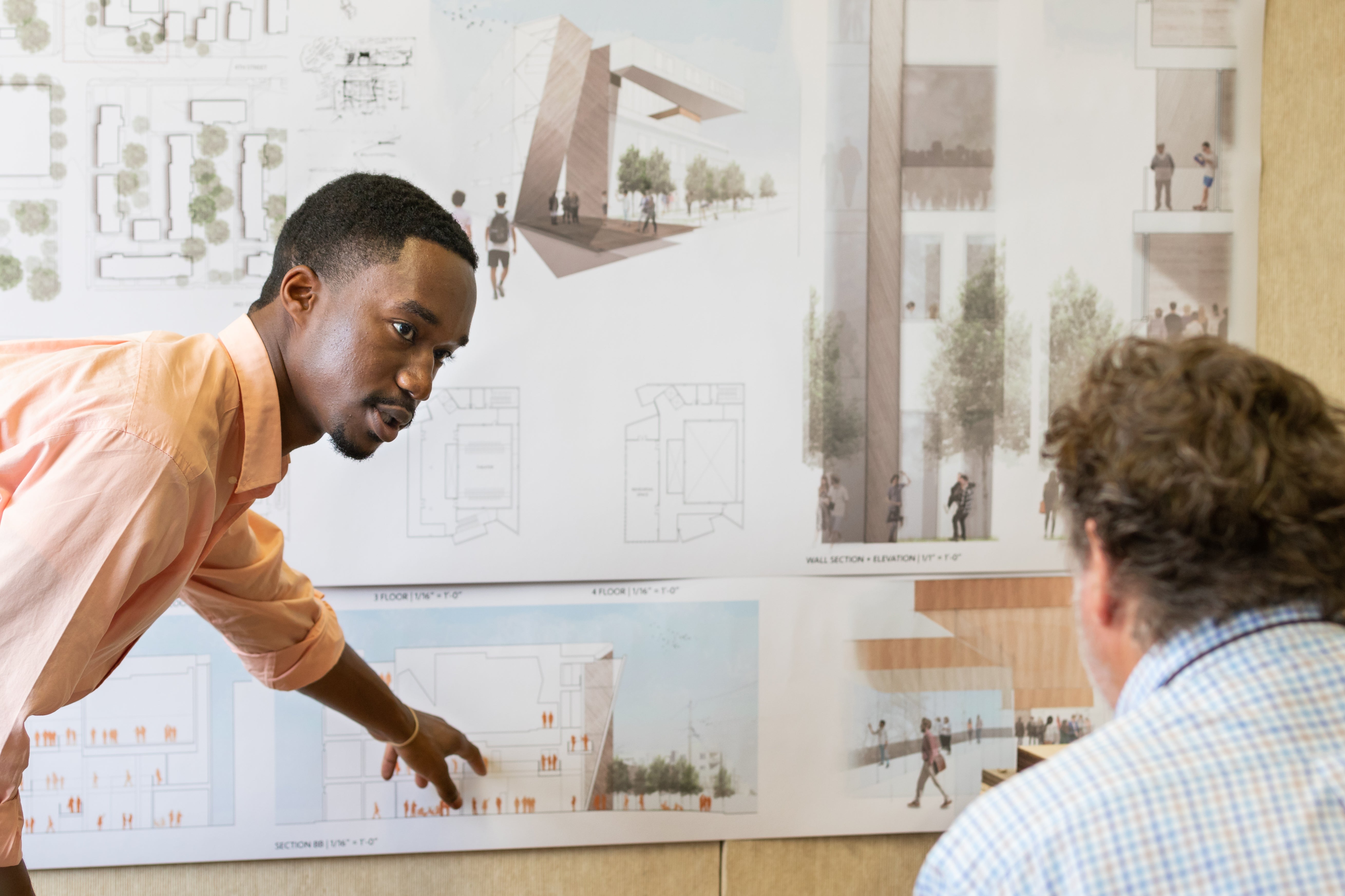 A student in a light orange shirt points at his pin up on the wall with a faculty member looking on