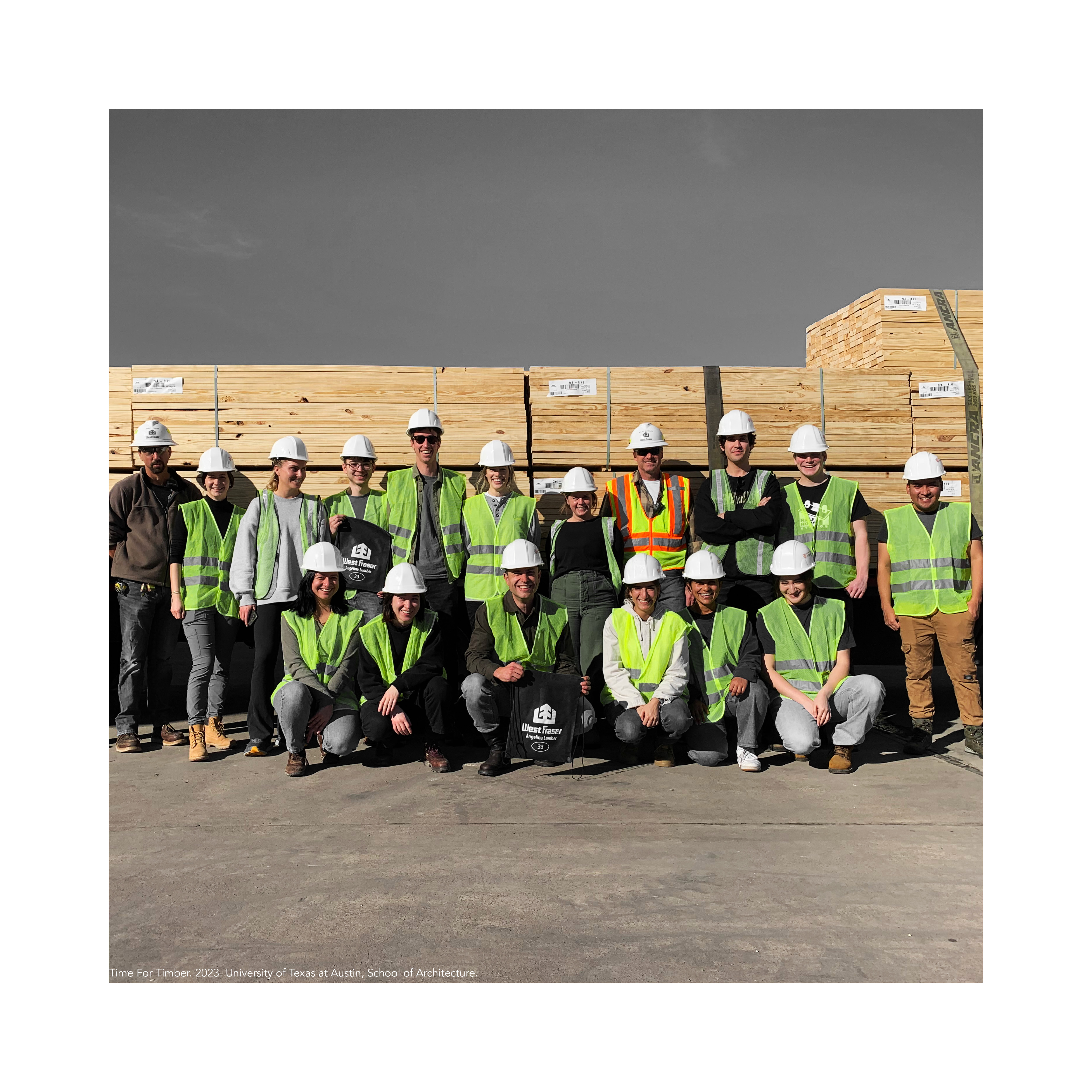 Students in neon green construction vests and hard hats posing as a group in front of tall piles of lumber.