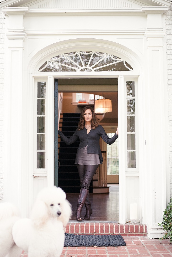 Lauren Rottet stands at the front door of a white house with a brick patio and a white poodle in the foreground.