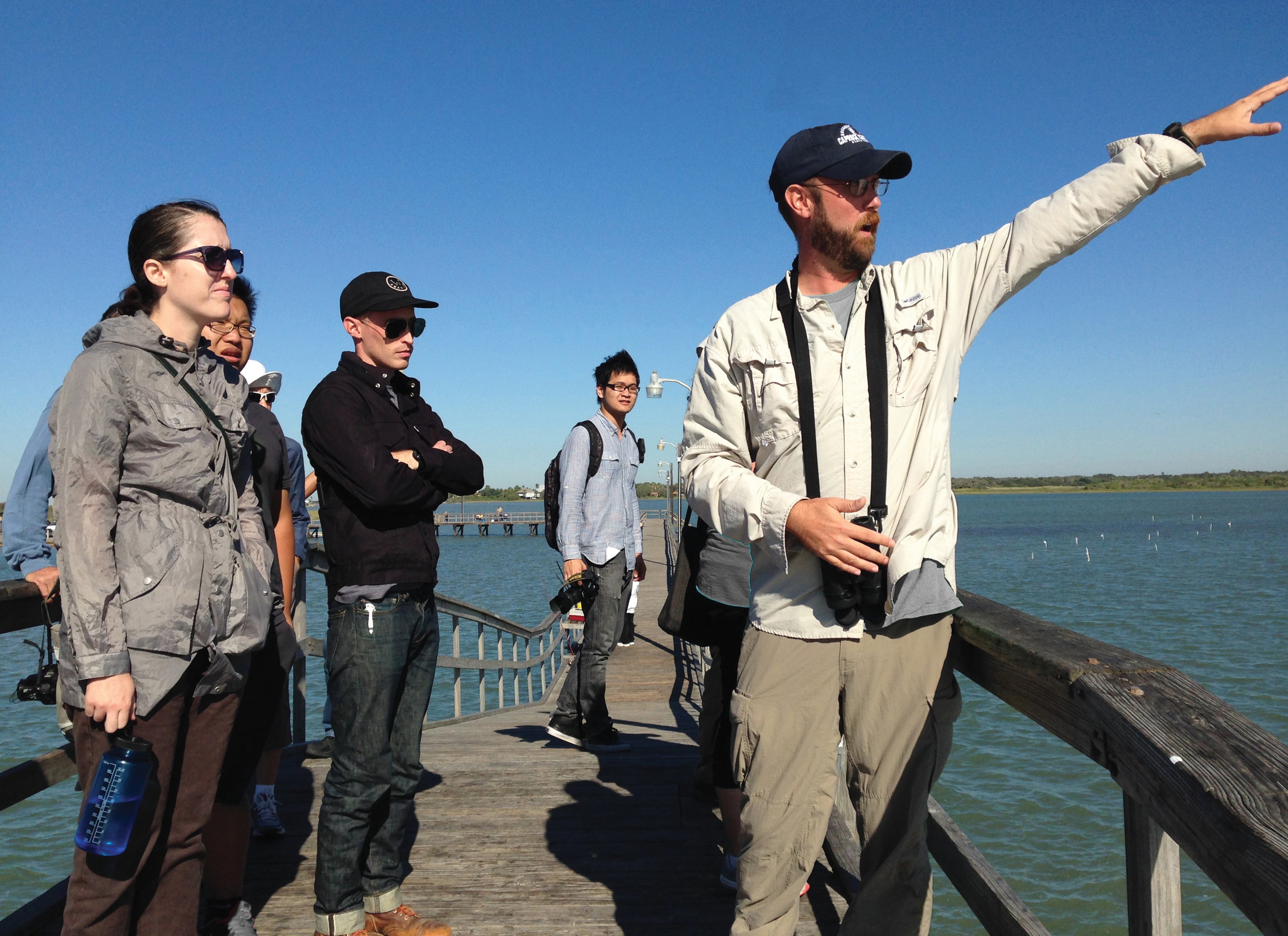 Several people standing on the bow of a boat looking off to the right of the camera as a man points into the distance.