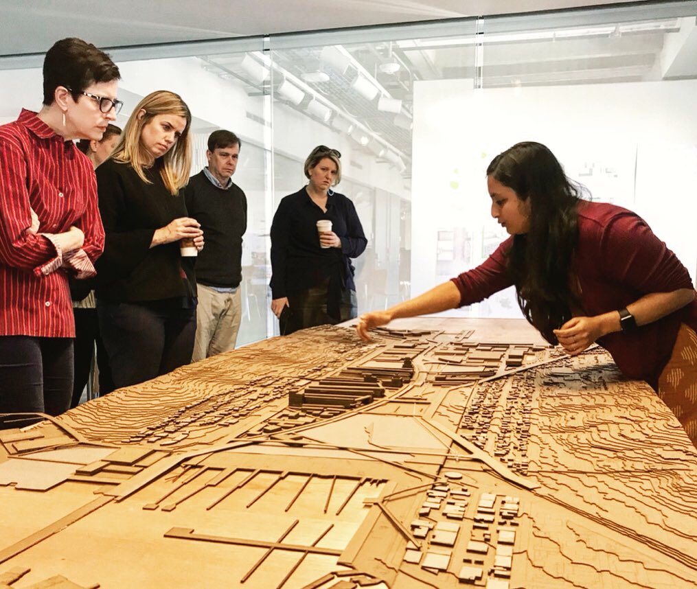Student leaning over and pointing to a physical model of a city, in front of a handful of reviewers