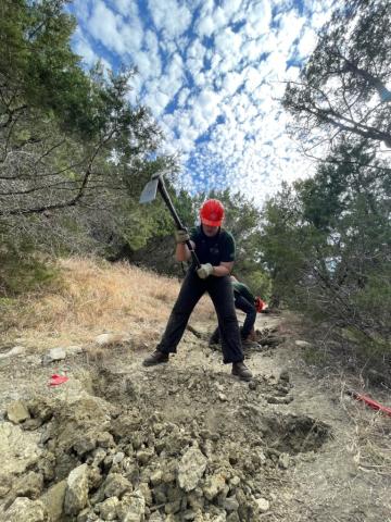 A person wearing a hard hat in mid-swing as they're breaking up the ground in a wooded area.