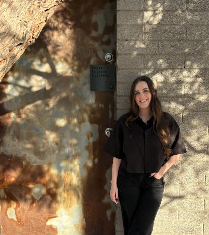 A young woman with brown hair leans against a brick wall. She is wearing all black.