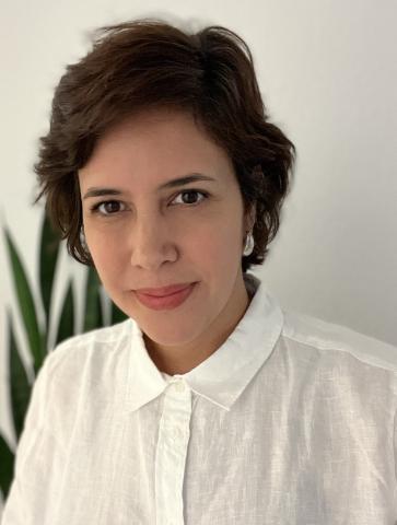 Luciana smiles at the camera against a white wall with a plant behind her