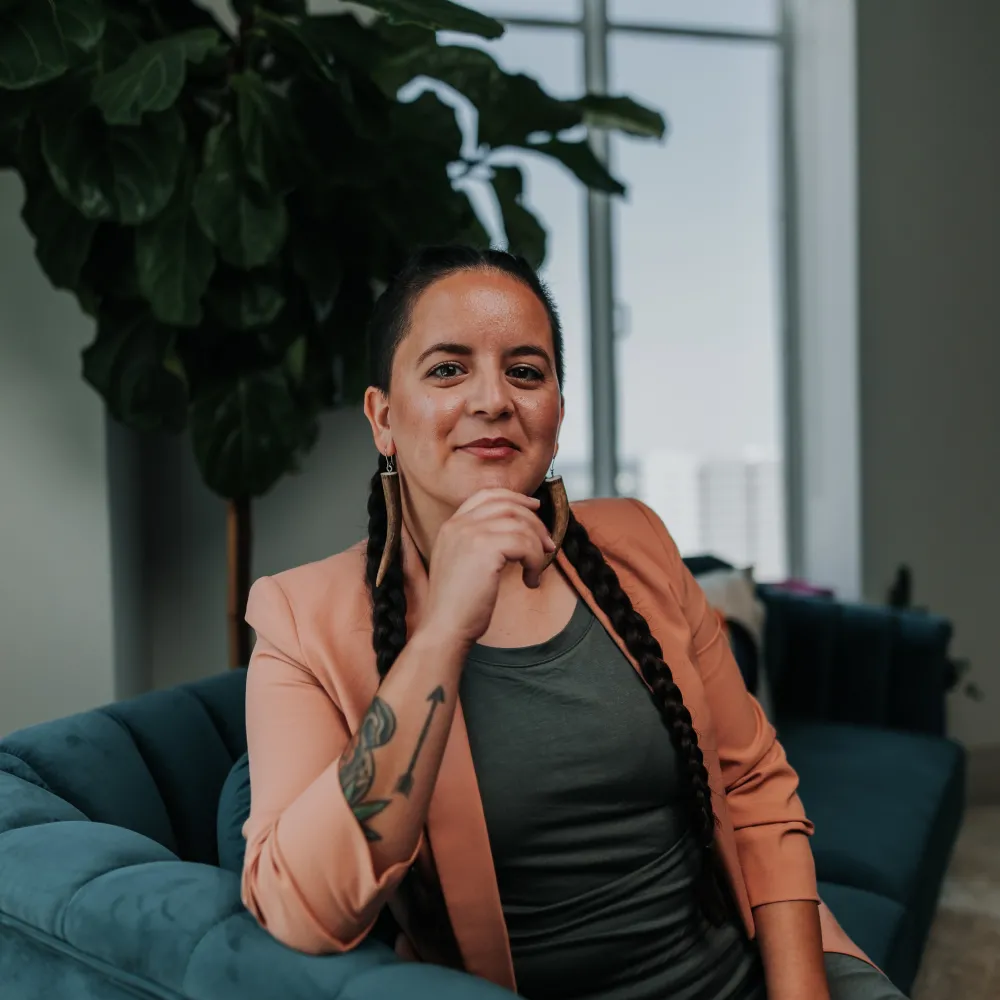  This is a picture of Emi Aguilar of the Indigenous Cultures Institute. Emi is seated on a blue couch in front of a tree and window in an interior setting.