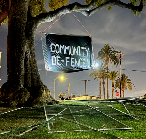 Sign "Community De-Fence!" hanging from a tree by a fallen fence