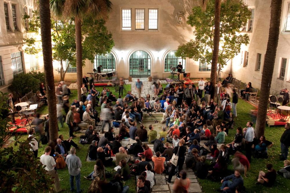 A large group of people gather in a courtyard at night, sitting on benches and grass, talking and socializing. Trees and arched windows surround the area, creating a lively, festive atmosphere.