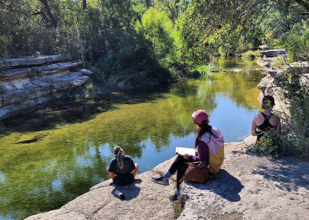 Three students sit on a cliff overlooking a watering hole with lush vegetation in the background