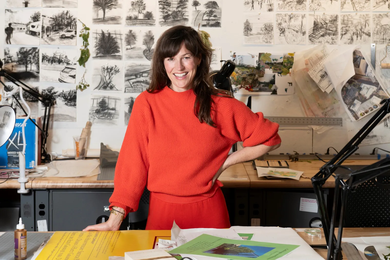 Sara Hendren, in red, standing in front of a desk with papers and materials on it, with drawings pinned up on the wall behind her