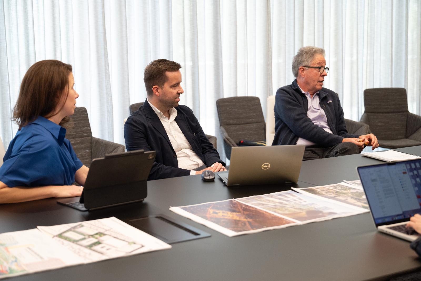 Three people sit at a desk and review project proposals. One is a woman with dark brown hair wearing a blue dress. Two are men wearing button up shirts and blazers. 