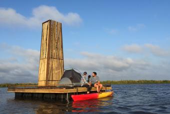Floating structure in the middle of a body of water with a tent on top and two people with a kayak
