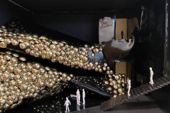 Design model with tiny figurines in a dark room with dozens of gold balls in an artistic display on the wall