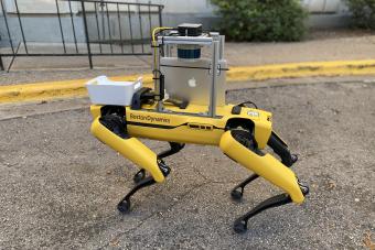 A yellow four-legged robot with an apple ipad attached stands on the sidewalk, poised to deliver cookies on campus