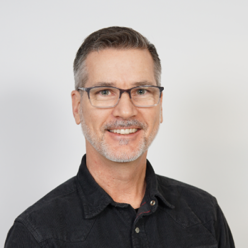 Headshot of Jason Radcliff; he is wearing glasses and a dark colored button up shirt, standing in front of a white background. He has dark grey hair.
