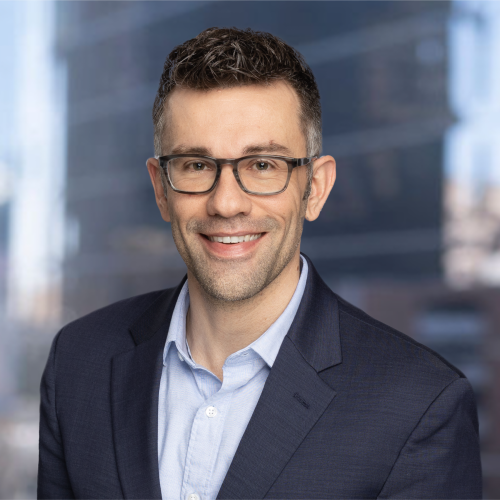 John Karras is smiling at the camera, wearing a blue collared shirt and navy blazer. He has dark rimmed glasses and brown hair. The background is blurred behind him.