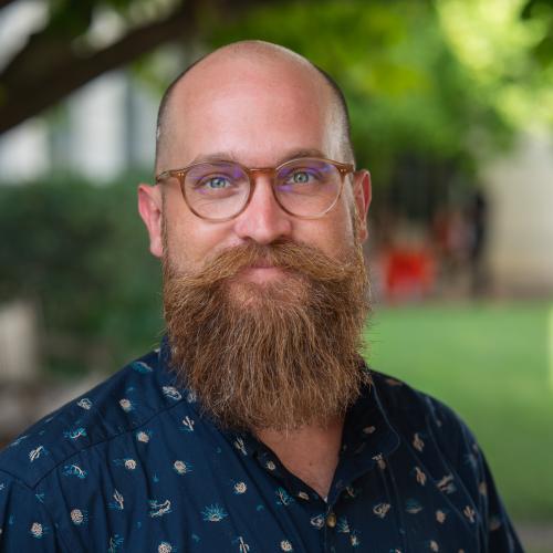 Daniel Scott is wearing a blue patterned collared shirt with brown rimmed glasses and a beard. He is smiling softly.