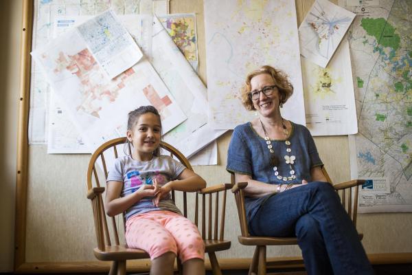 Liz Mueller sitting in front of a wall of maps and papers with a child in the chair next to her