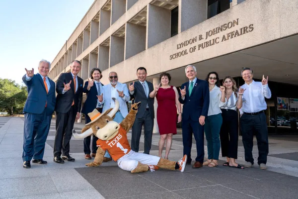 A group of people standing in front of the LBJ School of Public Affairs putting up the "hook em" sign with UT's mascot laying on the ground in front of them.