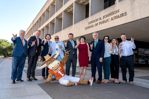 A group of people standing in front of the LBJ School of Public Affairs putting up the "hook em" sign with UT's mascot laying on the ground in front of them.