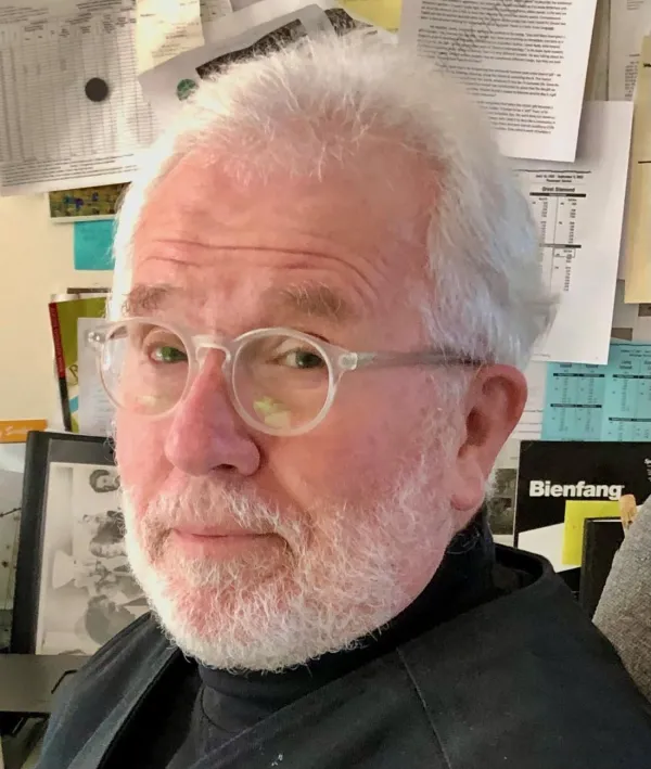 Steven A. Moore in a black turtleneck and clear glasses looking at the camera while standing in front of a wall of papers