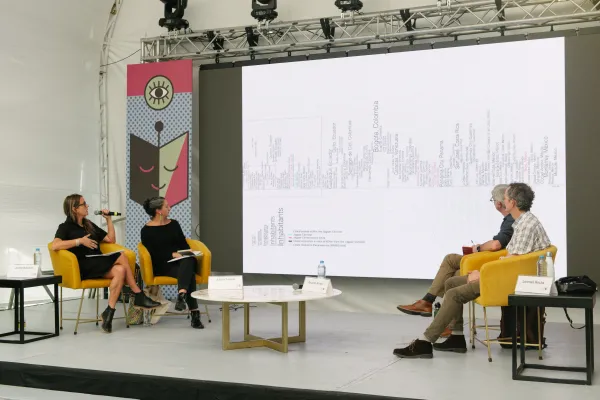Juana Salcedo and three other scholars sit on stage in yellow chairs in front of a projection screen.