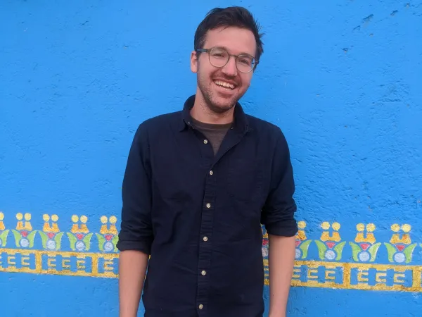 A man in glasses smiles in front of a bright blue wall.