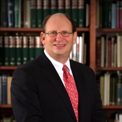 A man poses for a headshot wearing a suit and tie in front of a bookcase