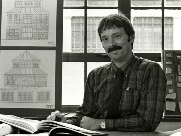 A photo of a young Professor Larry Speck, posing with architecture drawings behind him