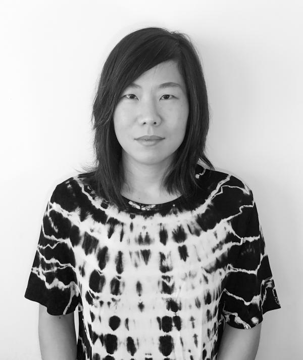 Black and white portrait of Stephanie Choi standing in front of a white wall in a tie dye top
