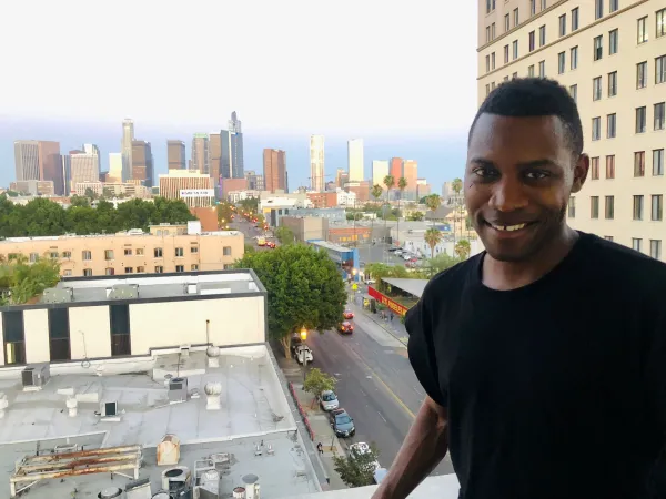 Tekena Koko smiles at the camera as he's standing on a balcony that overlooks an urban skyline at dusk