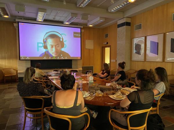 Seven people sit around an oval conference table in front of a projector screen, on which you can see a man in headphones speaking on a Zoom call