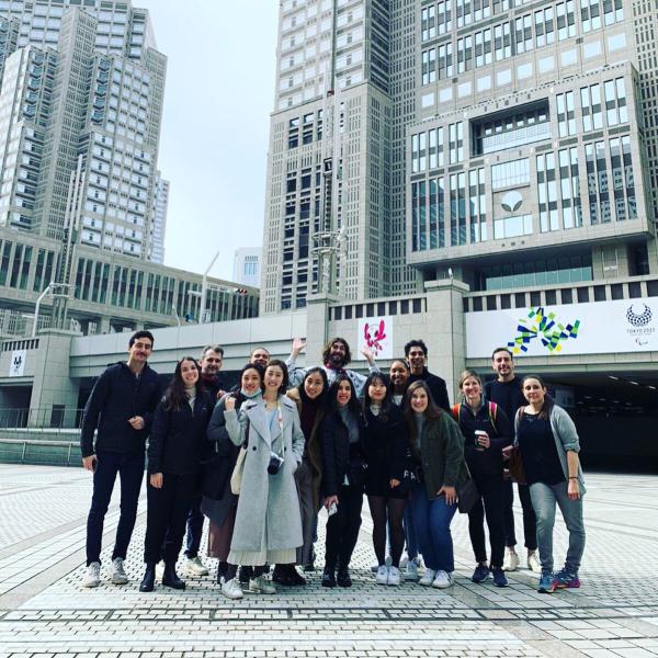A group of students and Adam Miller smile at the camera as a group, standing on a plaza in front of two office buildings in downtown Tokyo