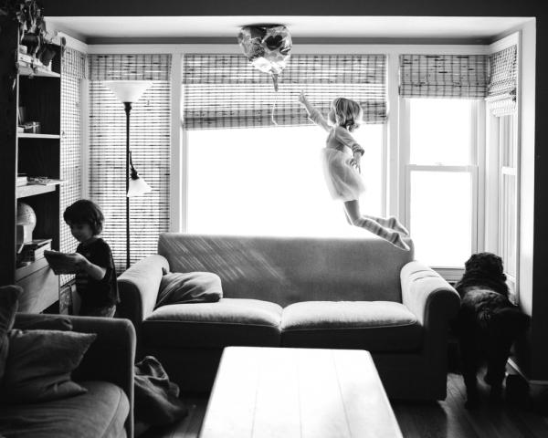 young girl mid-jump off sofa trying to reach a balloon that has floated to the ceiling