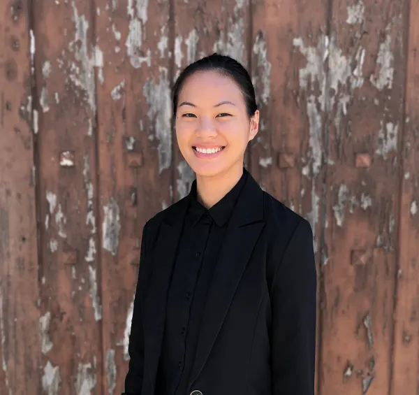 Xie Maggie Hill in all black smiling at the camera against a rustic wood background