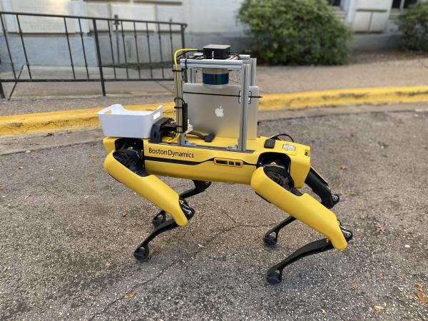 A yellow four-legged robot with an apple ipad attached stands on the sidewalk, poised to deliver cookies on campus