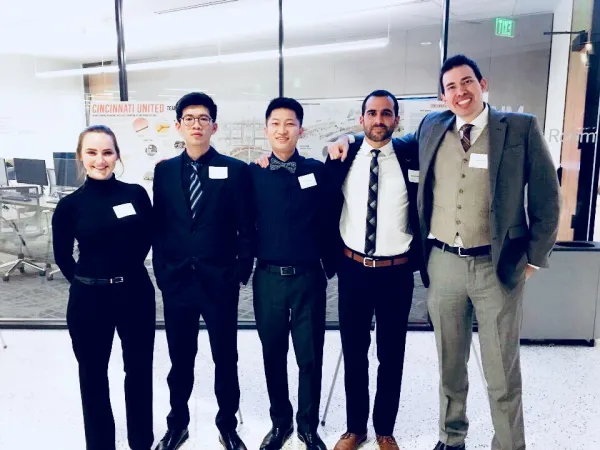 Five students smile at the camera in front of a floor-to-ceiling glass wall.