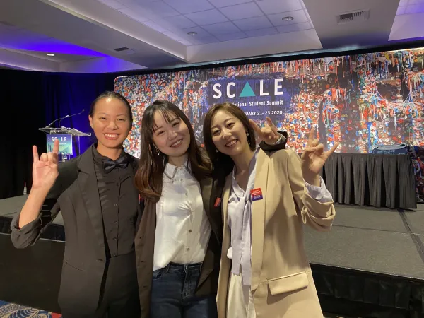 Three University of Texas at Austin interior design students smiling at the camera with their hands in the hook 'em symbol in front of a purple stage backdrop