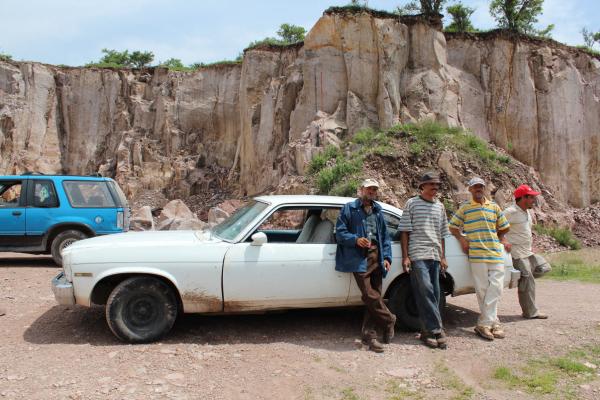 Four Mexican men in hats lean against a white sedan in front of a cliff of brown, rocky cantera stone