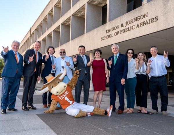 A group of people standing in front of the LBJ School of Public Affairs putting up the "hook em" sign with UT's mascot laying on the ground in front of them.