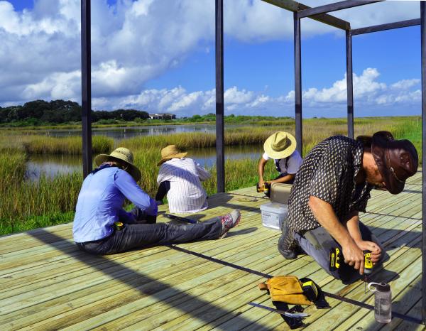 Several people kneeling and sitting as they work on a wooden platform that is surrounded by glass walls that overlook a marshy area and a blue sky