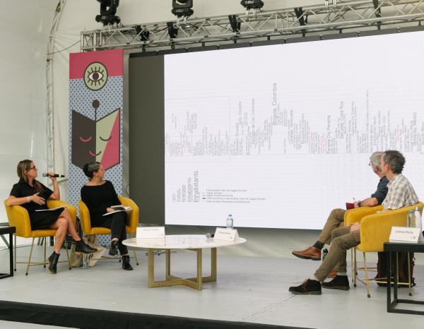 Juana Salcedo and three other scholars sit on stage in yellow chairs in front of a projection screen.