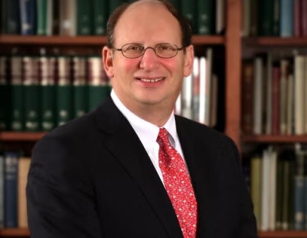 A man poses for a headshot wearing a suit and tie in front of a bookcase