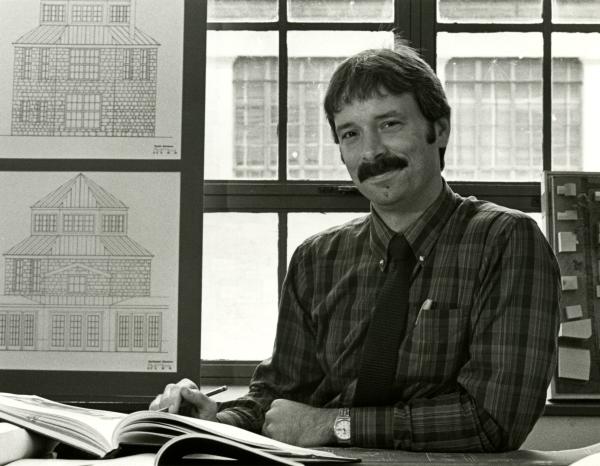 A photo of a young Professor Larry Speck, posing with architecture drawings behind him