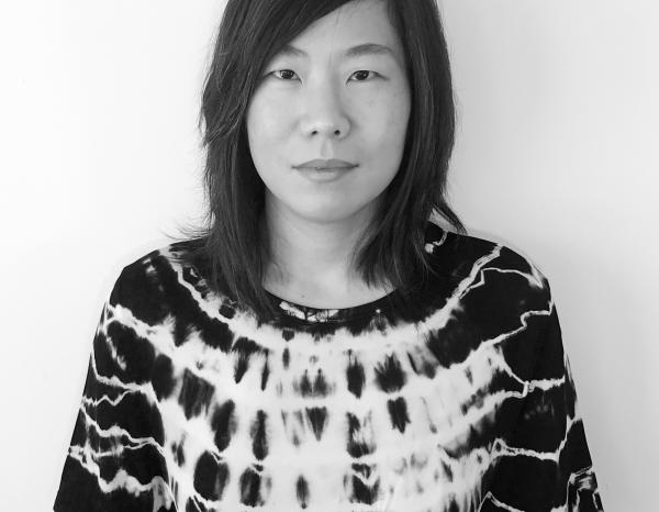 Black and white portrait of Stephanie Choi standing in front of a white wall in a tie dye top