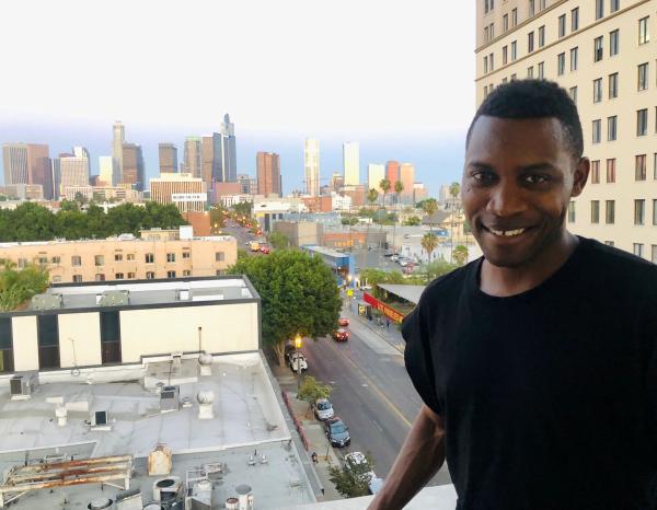 Tekena Koko smiles at the camera as he's standing on a balcony that overlooks an urban skyline at dusk