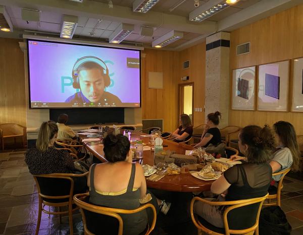 Seven people sit around an oval conference table in front of a projector screen, on which you can see a man in headphones speaking on a Zoom call