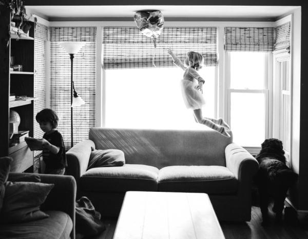 young girl mid-jump off sofa trying to reach a balloon that has floated to the ceiling